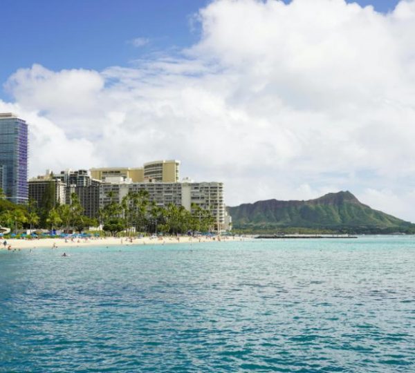 Türkisblaues Meer vor Waikiki Beach in Honolulu mit breitem Sandstrand, Hotels und dem markanten Diamond Head im Hintergrund.