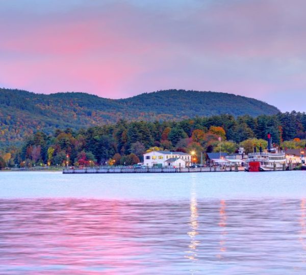 Ruhiges Wasser des Lake George mit bewaldeten Herbsthängen, Anlegern und rosa Abendhimmel auf einer Neuengland-Reise