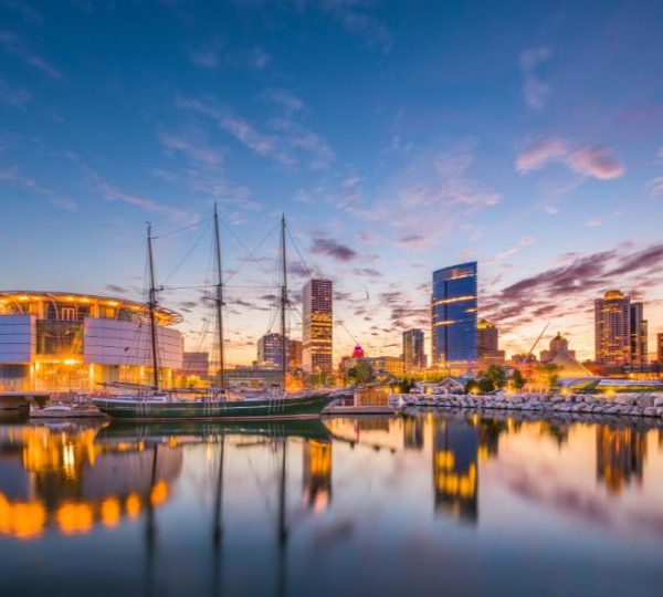 Abendliche Milwaukee-Skyline mit Segelschiffen am Wasser und beleuchteten Hochhäusern vor rosafarbenem Himmel