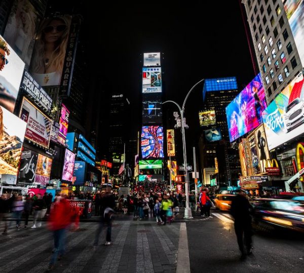 Menschen überqueren nachts eine belebte Kreuzung am Times Square in Manhattan zwischen leuchtenden Werbetafeln und Taxis.