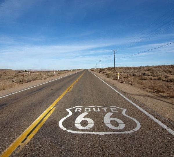 Gerade Landstraße mit gelber Mittellinie und weißem Route-66-Schild durch die karge Landschaft der östlichen Route 66