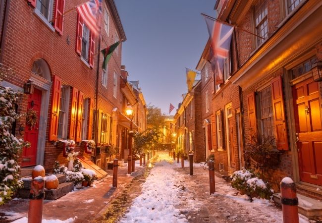 Verschneite Backsteinhäuser mit warm beleuchteten Türen säumen Elfreth’s Alley, die historische Wohnstraße in Philadelphia.