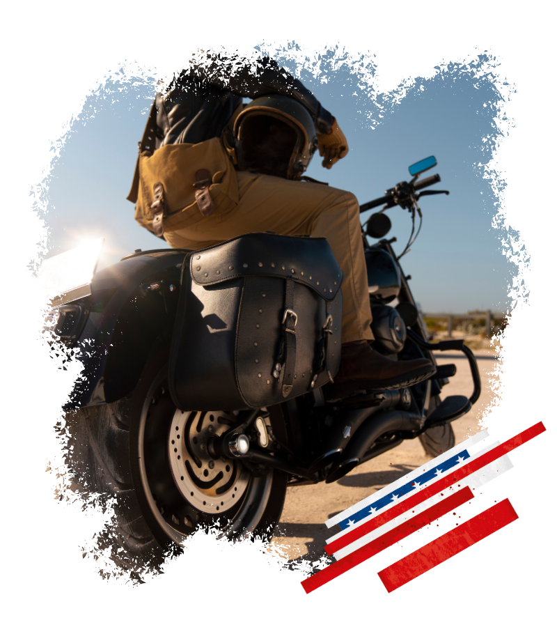 Ein Motorradfahrer sitzt auf einem schwarzen Cruiser vor blauem Himmel, daneben ein US-Flaggenmotiv in Rot, Weiß und Blau.