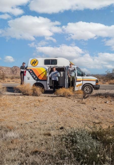Ein weißer Campervan mit bunten Streifen steht in einer trockenen Wüstenlandschaft, daneben stehen drei Reisende.