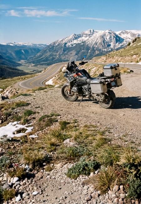 Ein beladenes Motorrad steht an einem Gebirgspass, während eine kurvige Straße ins Tal und schneebedeckte Gipfel führen.