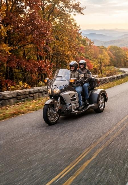 Zwei Personen fahren auf einem dreirädrigen Can-Am Spyder durch herbstliche Berge auf einer kurvigen Straße.