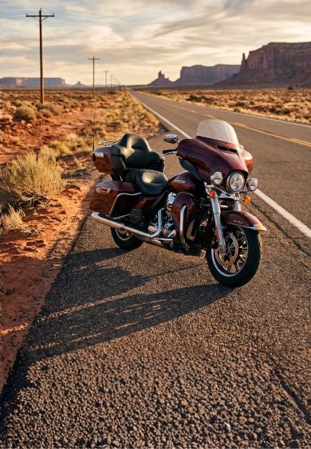 Auf einer langen Wüstenstraße steht ein maroonfarbenes Touring-Motorrad mit Verkleidung und Koffern neben rotem Felsland.