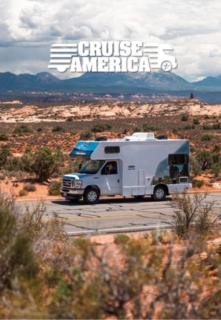 Ein weißes Cruise America Wohnmobil mit blauem Dach fährt durch eine trockene Wüstenlandschaft vor fernen Bergen.