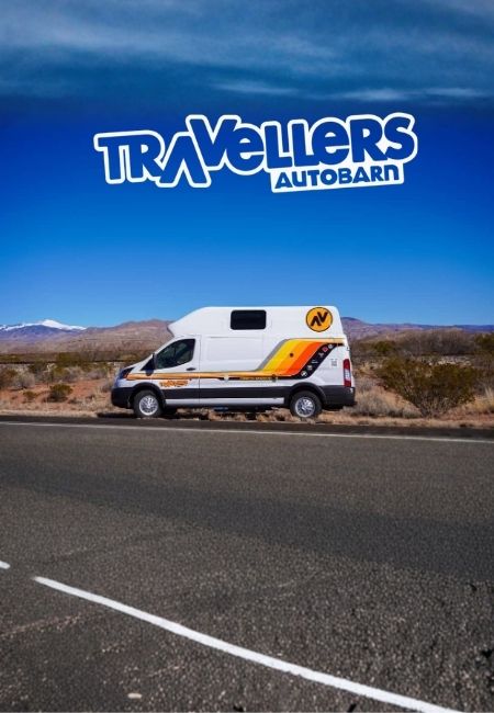 Ein weiß-orangefarbenes Wohnmobil von Travelers Autobarn steht an einer Straße vor weiter Wüstenlandschaft und Bergkulisse.