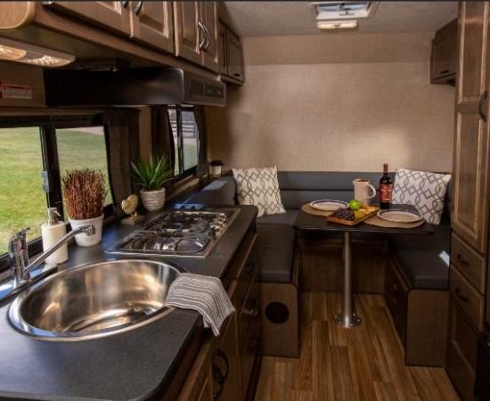 Der helle Innenraum eines Cruise America Compact Plus zeigt Küche, Spüle, Gasherd und eine kleine Sitzecke mit Tisch.