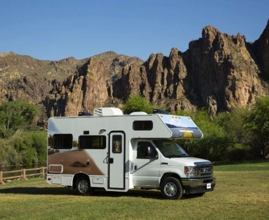Ein weißes Cruise America Wohnmobil steht auf einer Wiese vor steilen Felsformationen unter blauem Himmel.