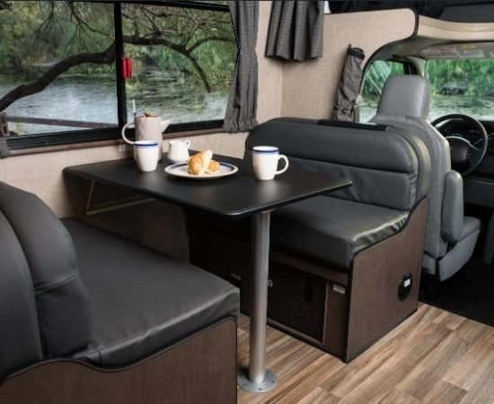 Im Wohnmobil-Innenraum stehen sich zwei Sitzbänke mit Esstisch gegenüber, dahinter sind Lenkrad und Fahrerplatz zu sehen.