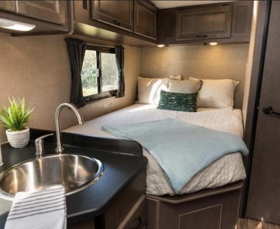 Im Cruise America Wohnmobil sieht man ein Bett neben der Küchenzeile mit Spüle, Fenster und dunklen Holzschränken.
