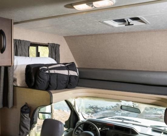 Im Cruise America 25 Standard liegt ein Bett über dem Fahrerhaus, daneben sind Sitzbank, Fenster und Stauraum zu sehen.