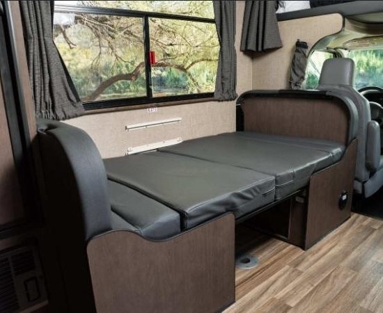 Im Cruise America Wohnmobil liegt ein schmaler Schlafbereich mit grauer Matratze und dunkler Polsterung vor dem Fahrerhaus.