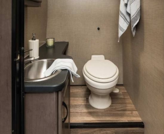 Im Cruise America Wohnmobil-Bad stehen eine weiße Toilette, ein kleines Waschbecken und ein Handtuch an beige verkleideten Wänden.
