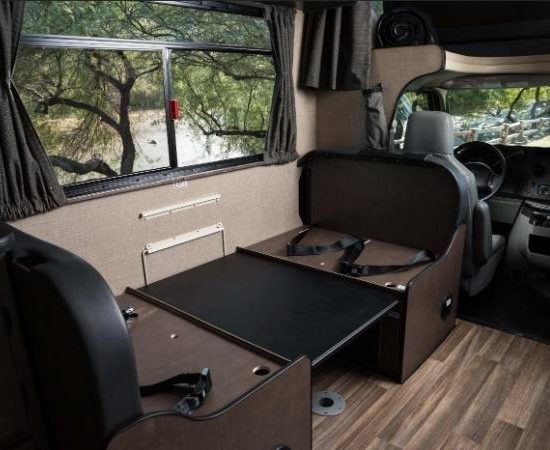 Im Cruise America Wohnmobil zeigt sich eine Sitzgruppe mit Tisch, großen Fenstern und Blick ins Grüne.
