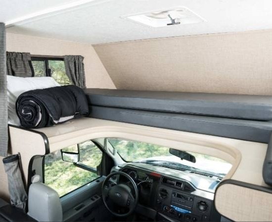 Das Alkovenbett mit grauen Matratzen und einer gerollten Decke liegt über dem Fahrerhaus im hellen Cruise America Wohnmobil.