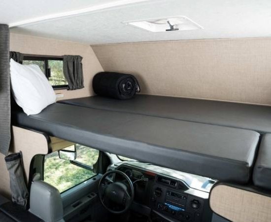 Im Cruise America Wohnmobil liegt eine graue Matratze über dem Fahrerhaus, daneben ein weißes Kissen und ein Fenster.