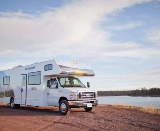 Ein weißes Cruise-America-Alkoven-Wohnmobil steht auf einer roten Uferstraße neben einem ruhigen See.