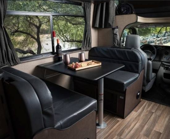 Im Cruise America Wohnmobil stehen eine Sitzgruppe mit Tisch, dunkle Polster und das Cockpit mit Blick nach vorn.