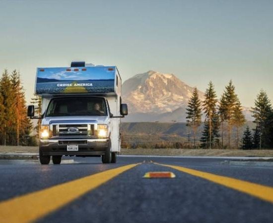Ein Cruise America Wohnmobil fährt auf einer Straße mit gelben Linien, dahinter stehen Bäume und ein hoher Berg im Licht.