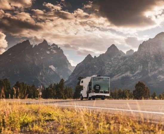 Ein Cruise America Wohnmobil fährt auf einer Straße vor steilen Bergen und dramatischen Wolken im warmen Abendlicht.