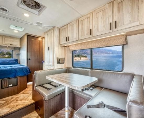 Im Road Bear Class C Wohnmobil stehen eine helle Sitzecke, eine Dinette und ein Schlafbereich mit Blick aufs Wasser.