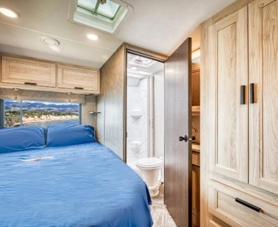 Im Road Bear Class C Wohnmobil zeigt sich ein Schlafzimmer mit blauem Bett, Holzschränken und offenem Badzugang.