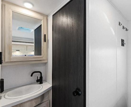 Im kompakten Bad des Road Bear Super Van Camper 23-25' stehen Waschbecken, Spiegel und dunkle Tür nebeneinander.