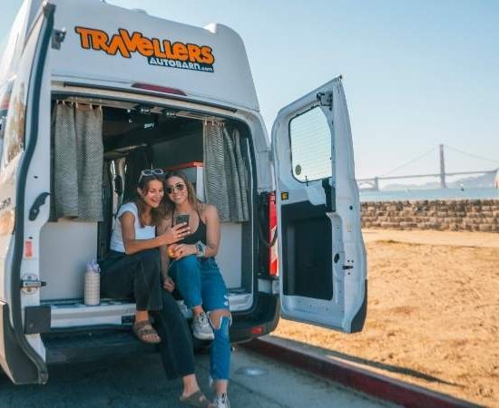 Zwei Reisende sitzen im geöffneten Travellers-Autobarn-Campervan an einer sonnigen Küstenstraße mit Blick auf eine Brücke.