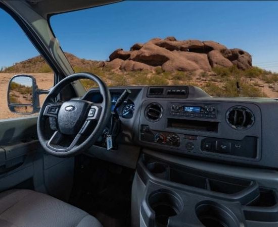Im Cockpit des Cruise America C18 blickt man durch die Windschutzscheibe auf eine staubige Wüstenstraße und große Felsen.