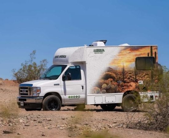 Ein weißer Cruise-America-Camper steht auf einer staubigen Piste in karger Wüstenlandschaft unter blauem Himmel.