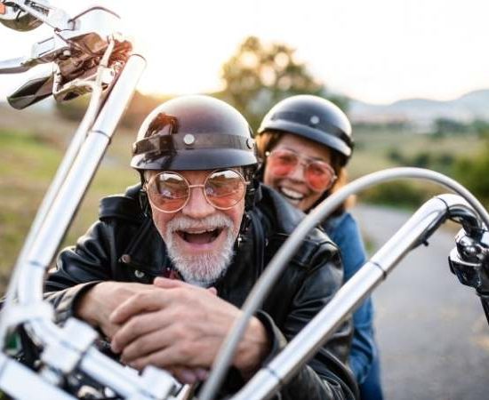 Ein lachendes älteres Paar fährt auf einem offenen Motorrad mit Helmen und Sonnenbrillen über eine Landstraße.
