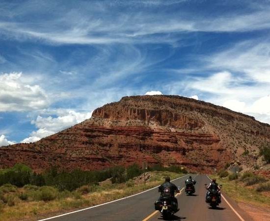 Drei Motorradfahrer fahren auf einer Asphaltstraße durch eine rote Felslandschaft unter weitem Himmel.