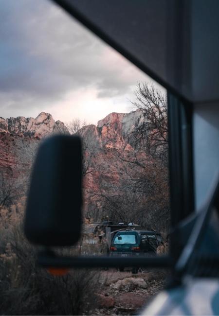 Durch das Wohnmobilfenster blickt man auf eine SUV und rote Felsen in einer kargen Berglandschaft.