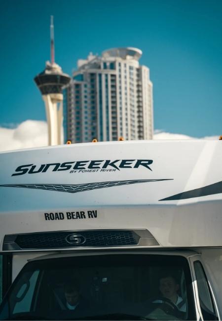 Ein weißes Road Bear RV Sunseeker mit sichtbarem Schriftzug steht vor einer Skyline mit markantem Turm.