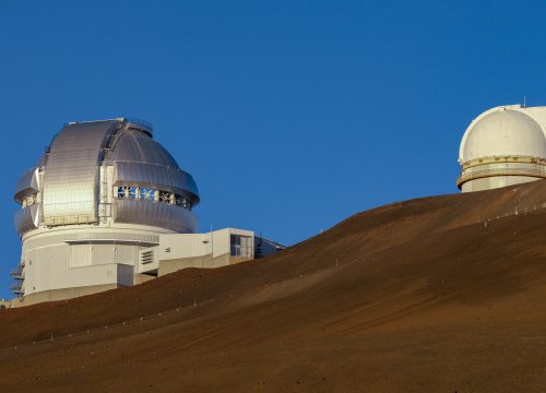 Mehrere astronomische Teleskope stehen am braunen Vulkanhang des Mauna Kea auf Big Island unter klarem blauem Himmel.