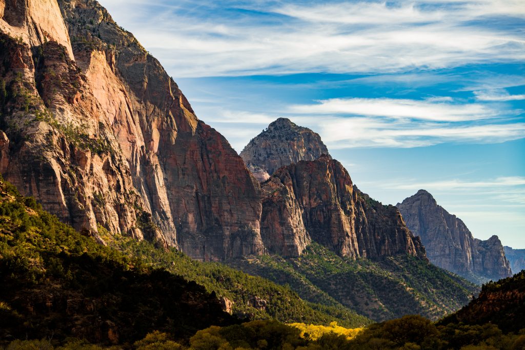 Rote Felswände und bewaldete Hänge prägen den tiefen Canyon des Zion National Park in Utah unter blauem Himmel.