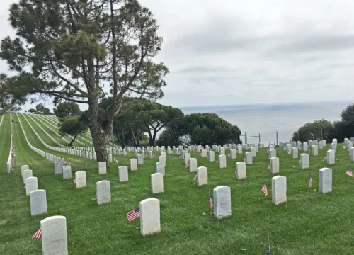 Geordnete weiße Grabsteine mit US-Flaggen stehen auf grüner Rasenfläche des Fort Rosecrans National Cemetery am Meer.