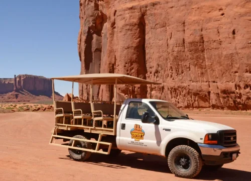 Ein offener Navajo-Tourtruck steht vor den roten Sandsteinfelsen des Monument Valley unter klarem, blauem Himmel.