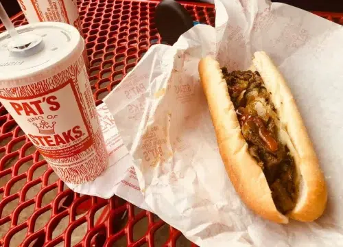 Auf einem roten Gittertisch liegt ein Philly Cheese Steak von Pat's King of Steaks neben einem Getränkebecher.