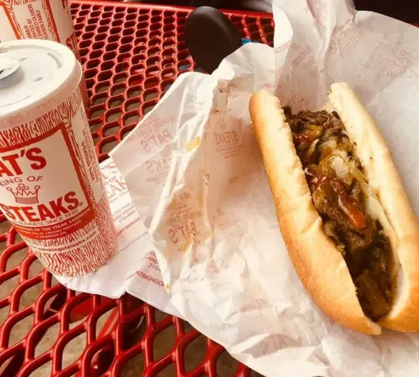 Auf einem roten Gittertisch liegt ein Philly Cheese Steak von Pat's King of Steaks neben einem Getränkebecher.