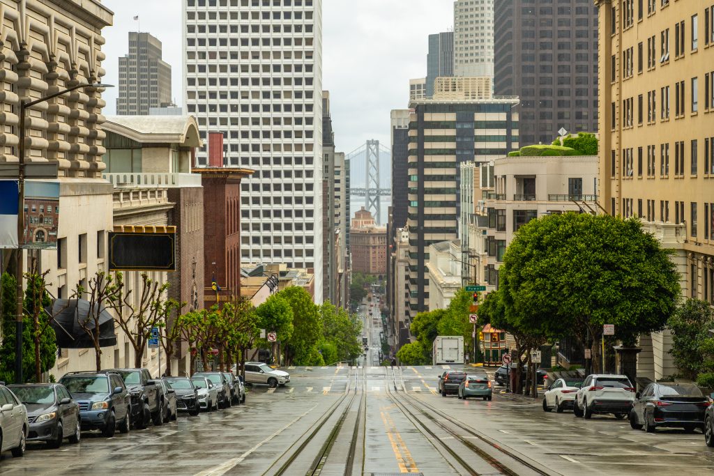 Nasse, steile Straße in San Francisco mit Cable-Car-Schienen, dichtem Stadtbild und der Bay Bridge im Hintergrund