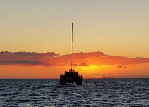 Ein Segelkatamaran fährt bei orangefarbenem Sonnenuntergang vor der Na Pali Coast auf Kauai über das offene Meer.