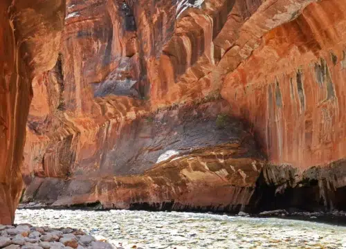 Wasserlauf zwischen den roten, meterhohen Felswänden der Narrows im Zion National Park mit sandfarbenem Gestein