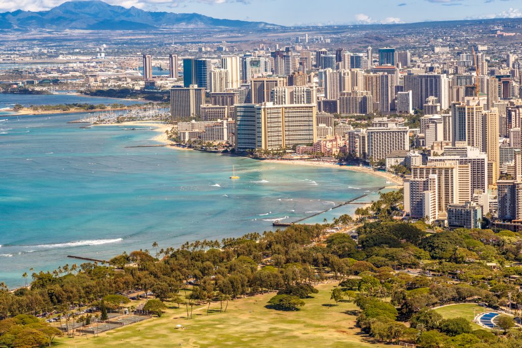 Türkisfarbene Küste von Waikiki Beach vor Honolulus Skyline, mit Diamond Head und grünen Parkflächen im Vordergrund.