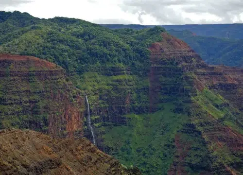 Rotes Gestein und grüne Hänge prägen den Waimea Canyon auf Kauai, während ein schmaler Wasserfall in die Schlucht stürzt.