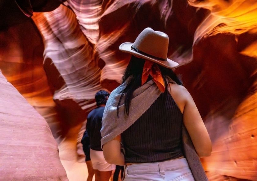 Eine Besucherin mit hellem Hut steht zwischen den rot-orange gewellten Sandsteinwänden des Antelope Canyon und blickt in die enge Schlucht.